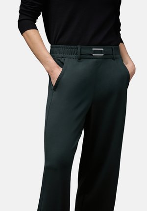 Pantaloni verdi scuri con una texture liscia, cintura elastica e un accento metallico sulla cintura. Presentano tasche laterali e una vestibilità sartoriale.