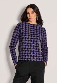 Vrouw in een paars en zwart houndstooth shirt met lange mouwen, met zwarte broek, staand met handen in de zakken tegen een effen achtergrond.