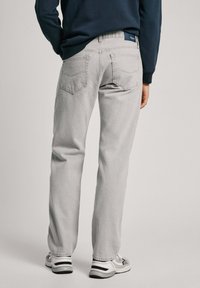 Gai pelēki džinsi ar taisnu piegriezumu, ar divām aizmugures kabatām un jostas etiķeti, valkāti ar melnām un baltām sporta kurpēm.