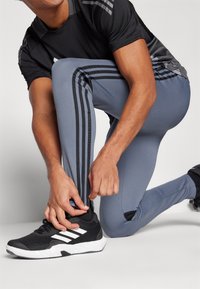 Sportstøj med sort kortærmet skjorte og grå tætsiddende bukser med sorte sideløsninger; model der justerer sorte sneakers med hvide detaljer.