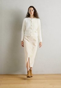 Cárdigan blanco de punto con botones, combinado con una falda midi crema con una abertura frontal. Botines marrones hasta el tobillo completan el conjunto.