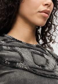 Γκρι υφασμάτινο τοπ με στρογγυλεμένο λαιμόκοψη, υφασματικές λεπτομέρειες και ξεφτισμένα στοιχεία στην περιοχή του στήθους. Το ύφασμα φαίνεται απαλό και ελαφρώς γυαλιστερό.