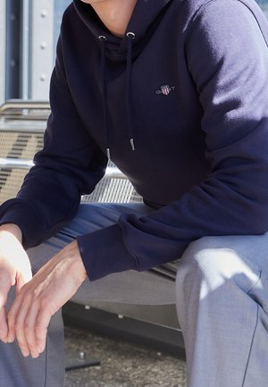 Osoba w granatowej bluzie z kapturem i jasnoszarych spodniach siedzi na metalowej ławce na zewnątrz, z rękami spoczywającymi na kolanach.