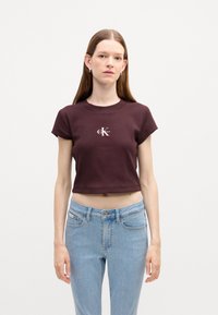 Bordowy skrócony t-shirt z krótkim rękawem i prążkowaną teksturą, z białym logo na środku. Noszony z jasnoniebieskimi dżinsami.