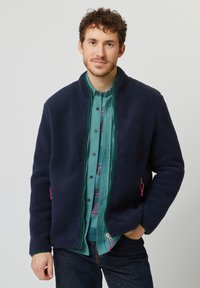 Marineblaue Fleecejacke mit hohem Kragen, Reißverschluss und seitlichen Taschen mit pinken Akzenten, getragen über einem türkisfarbenen Hemd.