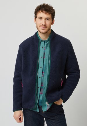 Navy fleece jacket with a high collar, zip closure, and side pockets featuring pink accents, worn over a teal button-up shirt.