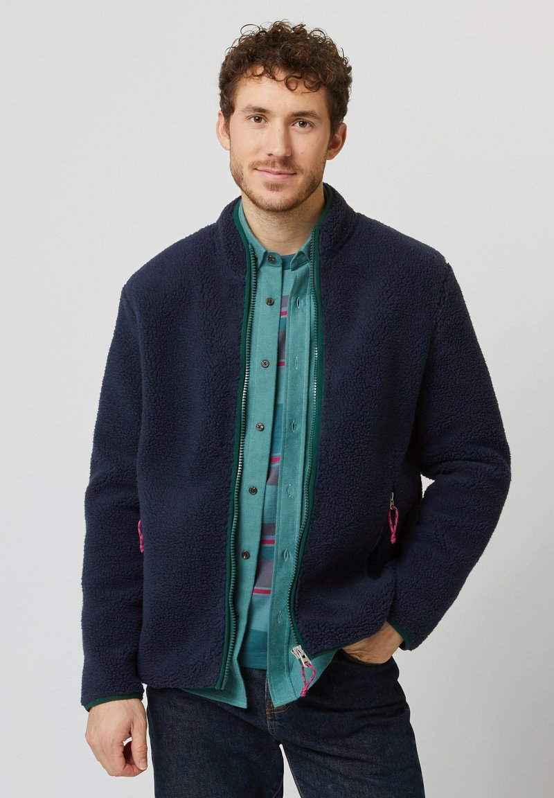 Marineblaue Fleecejacke mit hohem Kragen, Reißverschluss und seitlichen Taschen mit pinken Akzenten, getragen über einem türkisfarbenen Hemd.