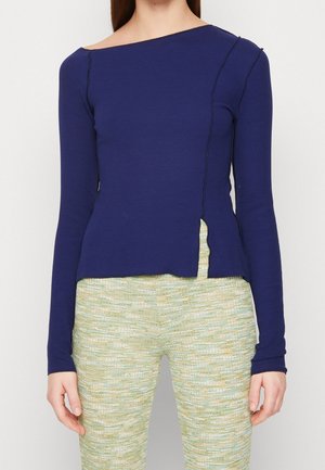 Femme vêtue d'un haut bleu marine ajusté à manches longues avec une fente asymétrique et d’un pantalon texturé vert clair sur un fond uni clair.