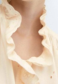 Blusa color pesca con colletto arricciato, dettagli in pizzo, tessuto morbido e una consistenza liscia, focalizzandosi su un design delicato e intricato.