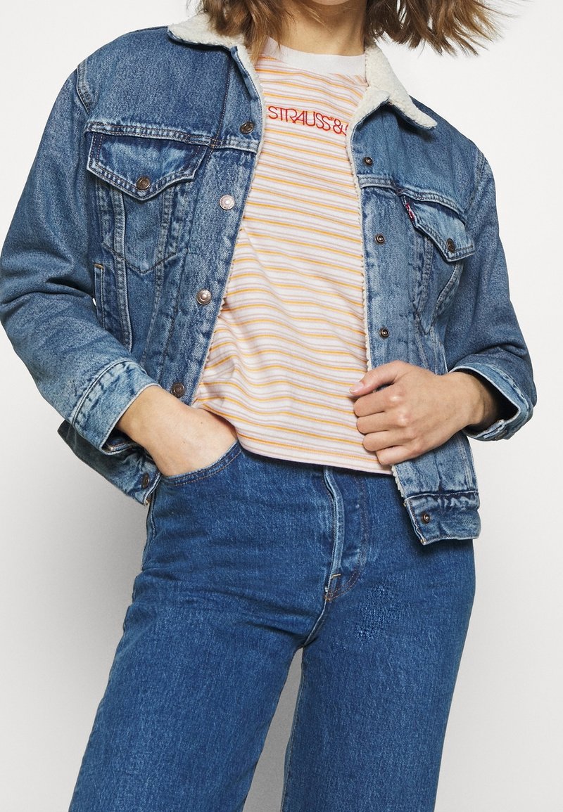 Giubbotto di denim con colletto in shearling, blu, con tasche con bottoni e dettagli usurati, indossato sopra una t-shirt a righe a maniche corte.