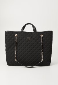 Sac cabas matelassé noir avec poignées courtes et longues lanières chaîne, orné d'une petite plaque logo triangulaire Guess au centre.