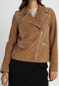 Hnědá suede motocyklová bunda se stříbrným kováním, asymetrickým předním zipem a několika zipovými kapsami. Má ramenní epauletky.