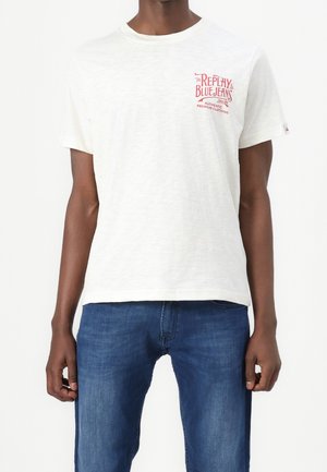 T-shirt blanc à manches courtes en tissu doux, avec un logo rouge imprimé sur le côté gauche de la poitrine. Associé à un jean en denim bleu.