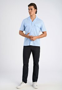 Vaaleansininen lyhyet hihansuut paita, jossa on kaksi etutaskua, napitettava malli ja rento istuvuus, yhdistettynä mustiin housuihin ja valkoisiin lenkkitossuihin.