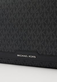 Svart håndveske med teksturert logo-mønster, en glatt lærdetalj og et sølvfarget "MICHAEL KORS"-logo merke nederst.