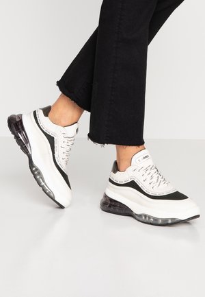 Person trägt schwarze Hose und weiße Sneaker mit schwarzen Akzenten und dicken transparenten Sohlen auf hellem Boden.