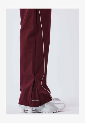 Pantaloni sportivi color granata con piping laterale bianco a contrasto, polsini con zip e logo "PXP" all'orlo. Abbinati a scarpe da ginnastica argentate.