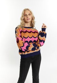 Maglione lavorato a maglia con un motivo a onde colorato in arancione, rosa e blu scuro, con polsini e orlo a costine, abbinato a pantaloni neri aderenti.
