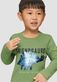 Grünes Langarmshirt mit einem blauen und silbernen Pailletten-Dinosaurierdesign. Der Text "FRIENDSAURS" steht über dem Dinosauriergrafik.