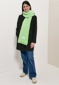 Manteau noir sur un haut blanc, associé à un jean bleu à jambes larges et des baskets marron clair. Une écharpe duveteuse vert clair avec des franges est portée autour du cou.