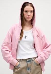 Felpa rosa chiaro con zip e tasca frontale, abbinata a una canotta bianca con un logo nero. Vestibilità morbida e tessuto soffice.