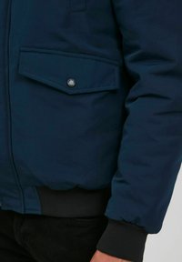 Navy blue padded jacket with a front flap pocket, silver button detail, elastic cuffs, and a smooth texture.