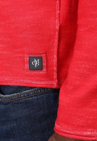 Rode gebreide trui met een klein zwart label met witte initialen aan de onderrand, gecombineerd met donkerblauwe jeans.