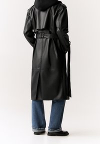 Trench-coat noir en cuir synthétique avec une ceinture à la taille, une fente haute à l'arrière et un design superposé. Associé à un jean bleu à jambes larges et des chaussures noires.