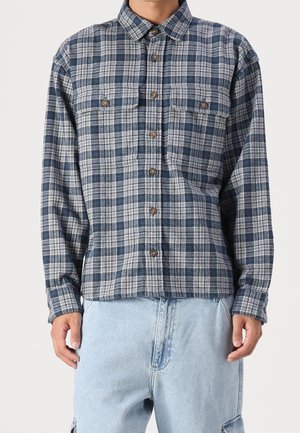 Mand iført blå og hvide ternet skjorte med brystlommer og lyseblå denim bukser, stående mod ensfarvet baggrund.