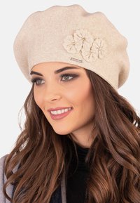 Berretto beige lavorato a maglia con tre applicazioni di fiori texture e accenti di perle. Il cappello ha un design largamente morbido e una texture a coste.