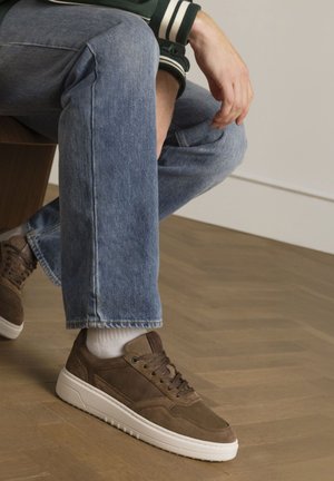 Bruine suède sneakers met een witte zool, met veters, een gladde afwerking en accenten. Draagt met lichtblauwe spijkerbroeken en witte sokken.