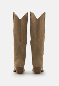 Beige Wildleder-Kniestrumpf-Stiefel mit spitzen Oberteilen, aufwendigen Nähmustern, seitlichen Reißverschlüssen und gestapelten Absätzen. Sichtbare strukturierte Oberflächendetail.