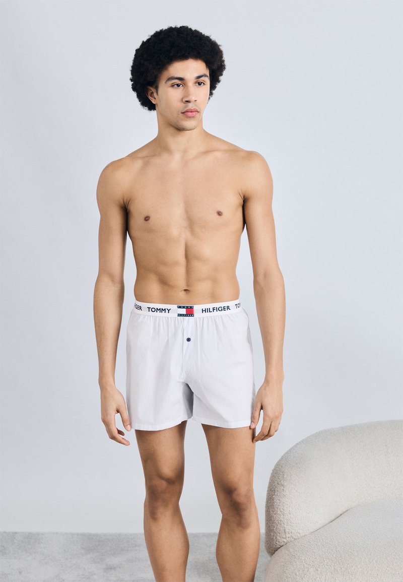 Λευκά βαμβακερά μποξεράκια με οριζόντιες ρίγες, με λάστιχο μέσης με λογότυπο και ένα κουμπί στο καβάλο. Το μοντέλο στέκεται μπροστά σε φωτεινό φόντο.