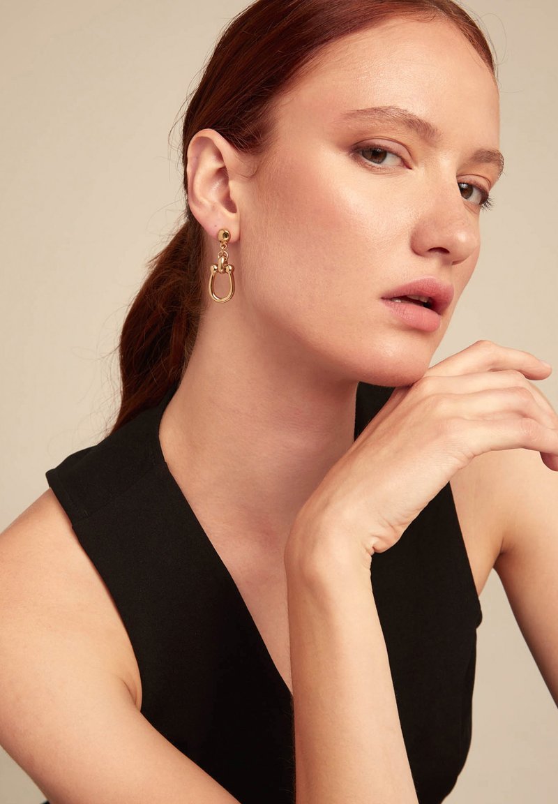 Pendientes de aro dorados con un Stud redondo y detalles entrelazados. El diseño presenta metal liso y un acabado pulido.