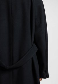 Cappotto di lana nera con una larga cintura annodata all'indietro, manica visibile con polsino abbottonato e una mano parzialmente visibile.