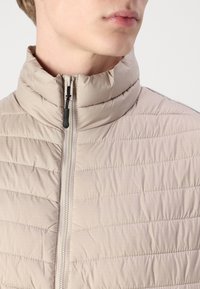 Light beige puffer jacket with a high collar, diagonal quilted design, and front zipper. Material has a smooth, slightly shiny texture.