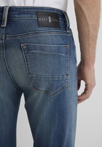 Jeans in denim blu scuro con un lavaggio sbiadito, caratterizzati da un'etichetta in pelle nera sulla vita posteriore e una singola tasca posteriore con dettagli di cucitura.
