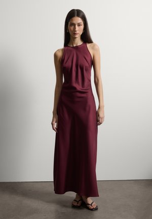 Frau mit langen dunklen Haaren, die ein ärmelloses, bordeauxfarbenes Maxikleid aus Satin und schwarze Sandalen trägt, steht vor einer schlichten grauen Wand.