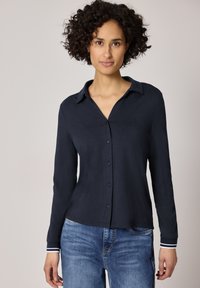 Femme aux cheveux courts et bouclés portant une chemise boutonnée à manches longues de couleur bleu marine foncé et un jean bleu, posant devant un fond uni.
