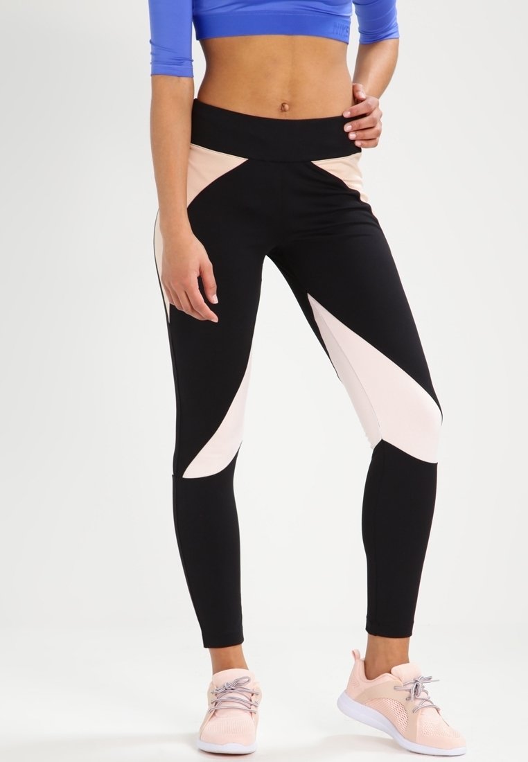 Frau in schwarz-beigen, farbblockierten Leggings, einem blauen Crop-Top und hellrosa Sportschuhen, steht vor einem weißen Hintergrund.