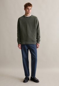 Zielony sweter z prążkowanymi mankietami i dołem, noszony na białej koszuli, w połączeniu z ciemnoniebieskimi spodniami i czarnymi butami, stojący na neutralnym tle.