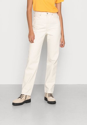 Person iført hvide bukser med lige ben, beige snørestøvler med sorte såler og en gul top med korte ærmer, stående mod en ensfarvet baggrund.