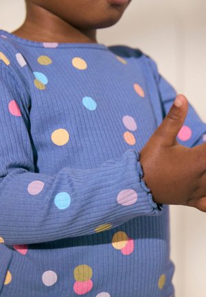 Child wearing a blue, long-sleeve shirt with multicolored polka dots, holding hands close to their chest against a plain background.