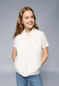 Biela vypchávkovaná vesta s ružovými akcentmi, prednými gombíkmi a bočnými vreckami. Nosená cez svetlú pletenú blúzku s krátkym rukávom.