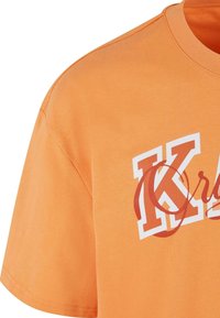 Orange bomulds t-shirt med et hvidt og orange grafisk print med det fremtrædende bogstav 'K'. Korte ærmer og blød tekstur.