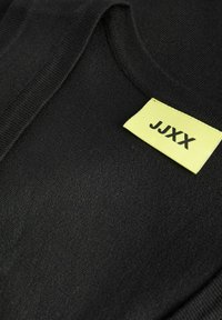 Μαύρο πλεκτό ένδυμα με στρογγυλή λαιμόκοψη και μια κίτρινη ορθογώνια ετικέτα ραμμένη κοντά στον γιακά με την επιγραφή "JJXX" σε μαύρο χρώμα.