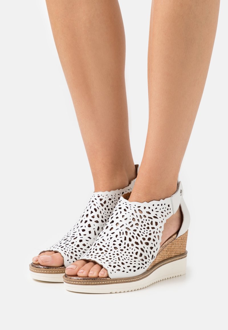 Pieds portant des sandales compensées en cuir blanc perforé, à bouts ouverts et brides arrière, sur une semelle plateforme blanche.