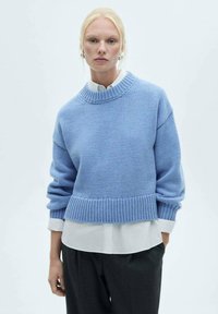 Hellblau gestrickter Pullover mit hohem, geripptem Halsausschnitt und verkürztem Design. Hat lockere Ärmel und einen gerippten Saum; wird über einem weißen Hemd mit Kragen getragen.