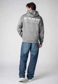 Gray hoodie with a large white text print on the back, paired with loose-fitting blue denim jeans and white sneakers.