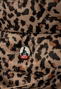 Beige gewatteerd jack met een zwart-bruine luipaardprint. Heeft een ronde patch met "JOTT" en een afbeelding van een scooter.